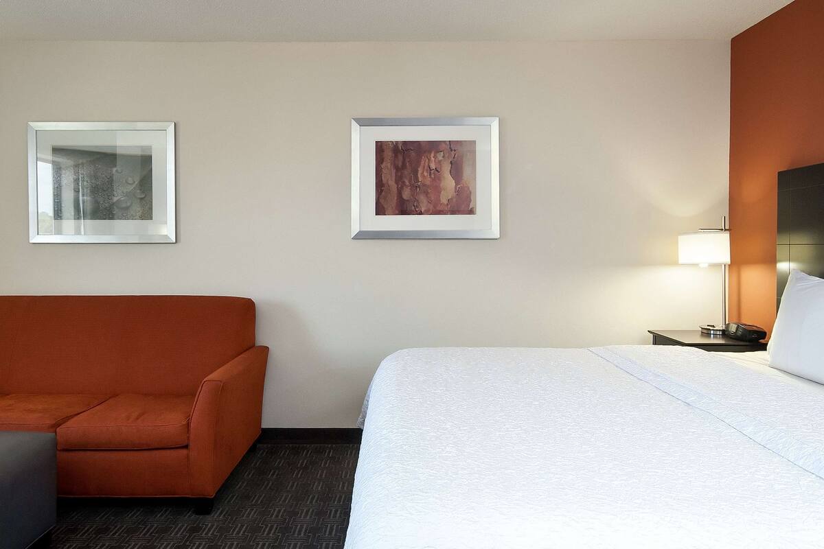 Una habitació d'hotel amb un llit, un sofà, dues fotografies emmarcades a la paret i una tauleta de nit amb una làmpada.