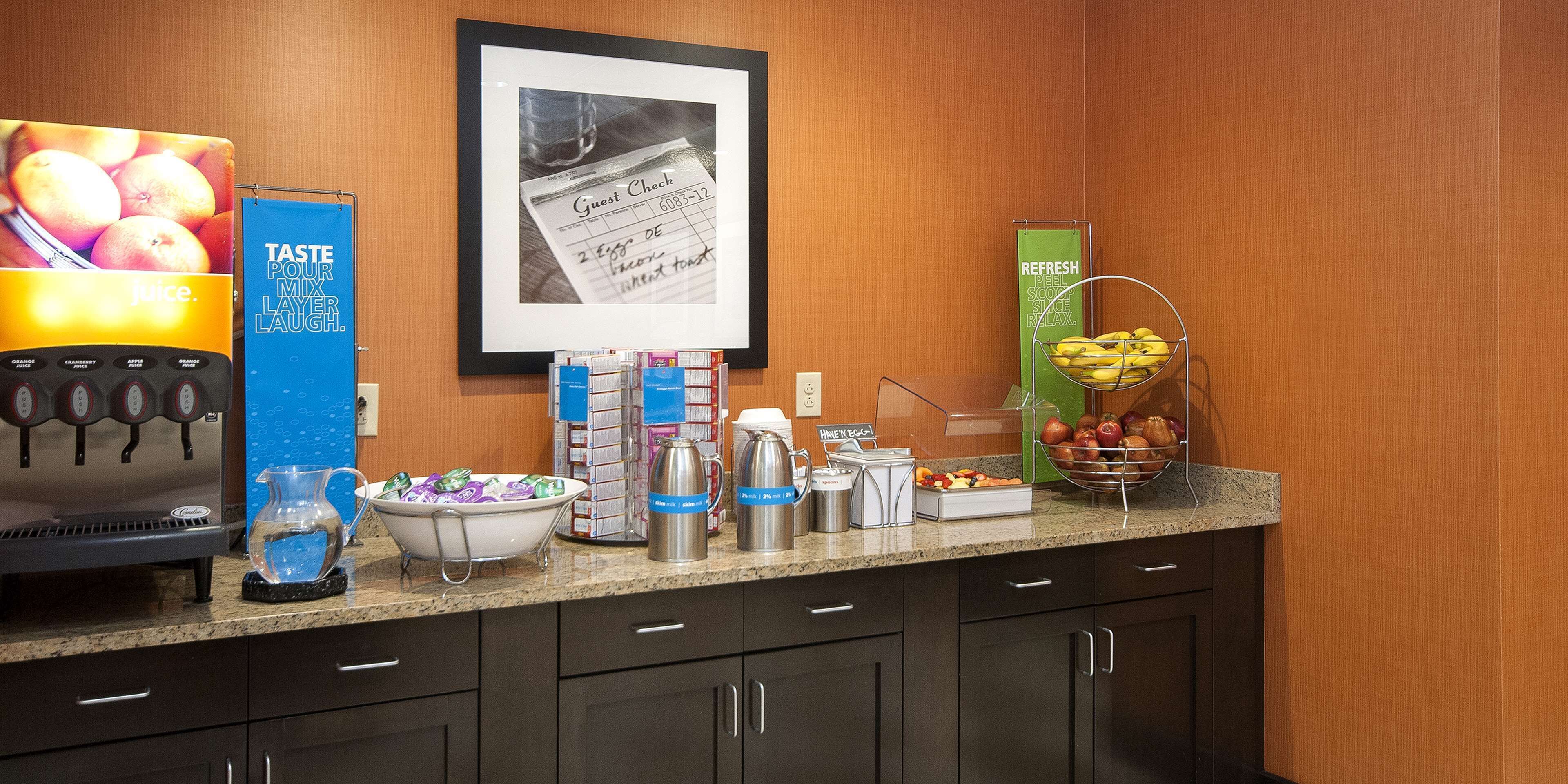 Un espace où est servi le petit-déjeuner à l’hôtel, avec une machine à café, un distributeur de jus et divers aliments disposés sur le comptoir.