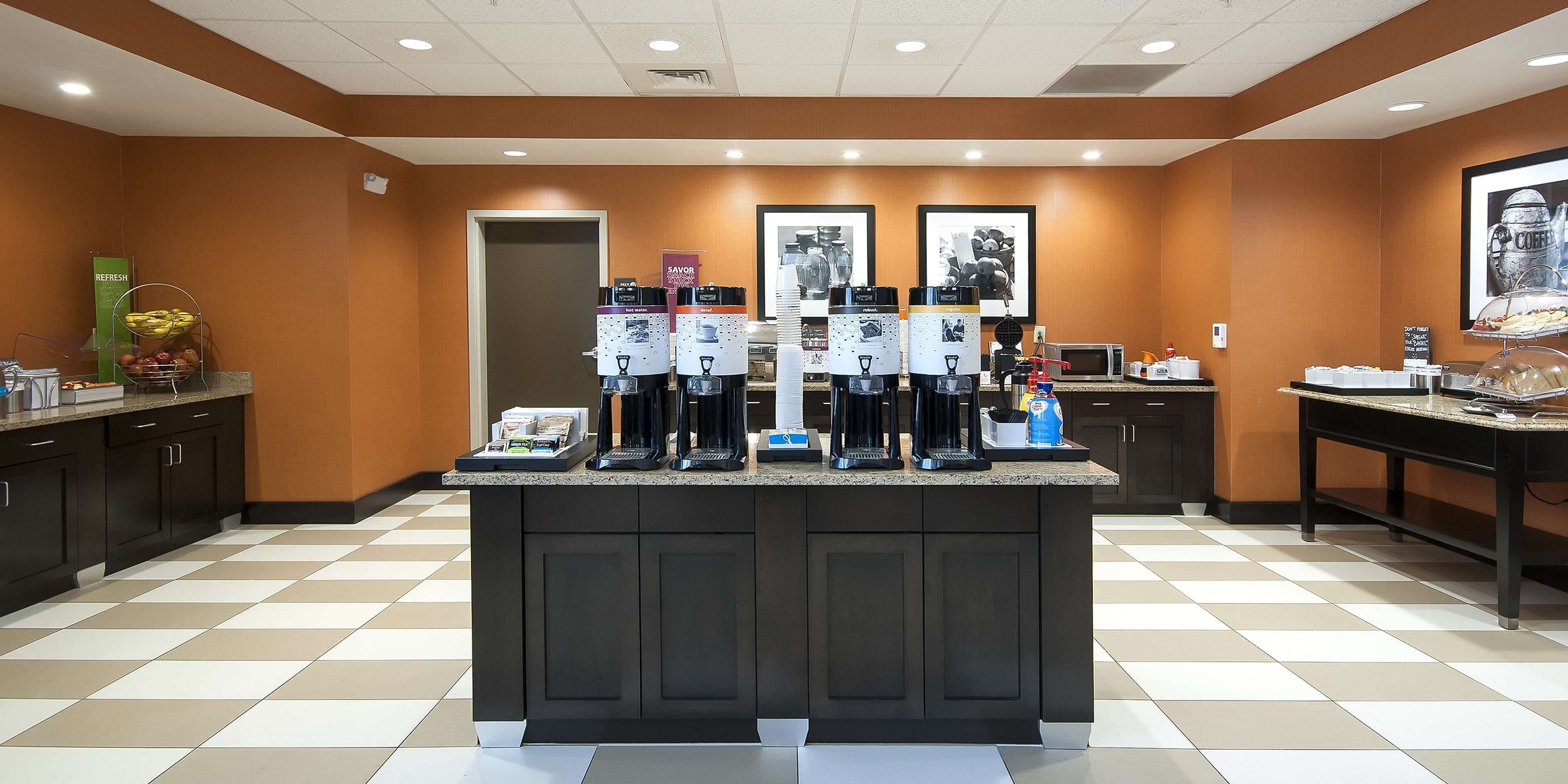 A hotel kitchen with a central island featuring coffee makers, a fruit basket, and a microwave.