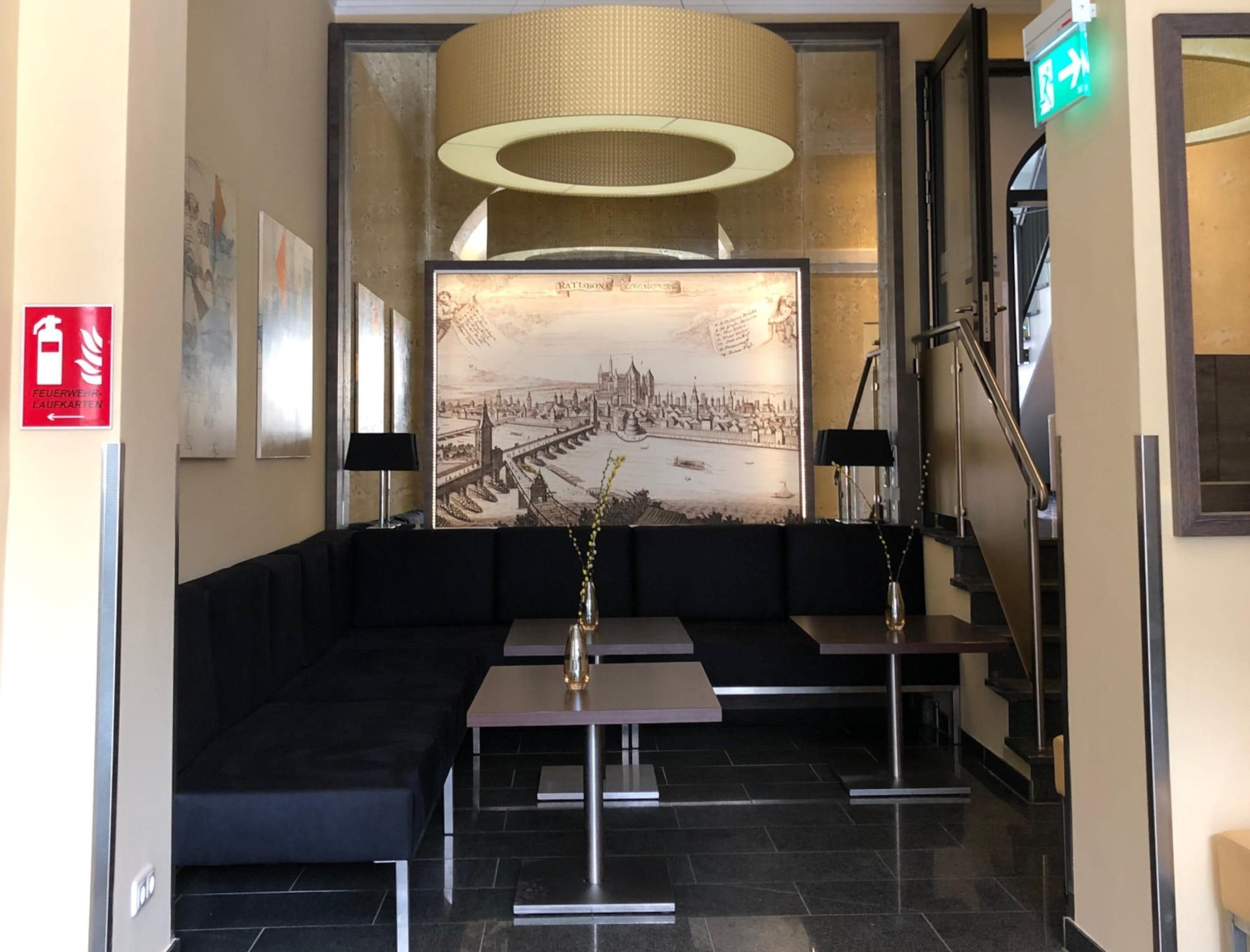 A lobby with a black sofa, two tables, a large framed cityscape artwork, a fire exit sign, and a staircase.
