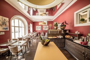 Un restaurant avec un plafond haut, des fenêtres en arc de cercle et des œuvres d’art accrochées aux murs. Les tables sont disposées avec des chaises, et un buffet est visible.
