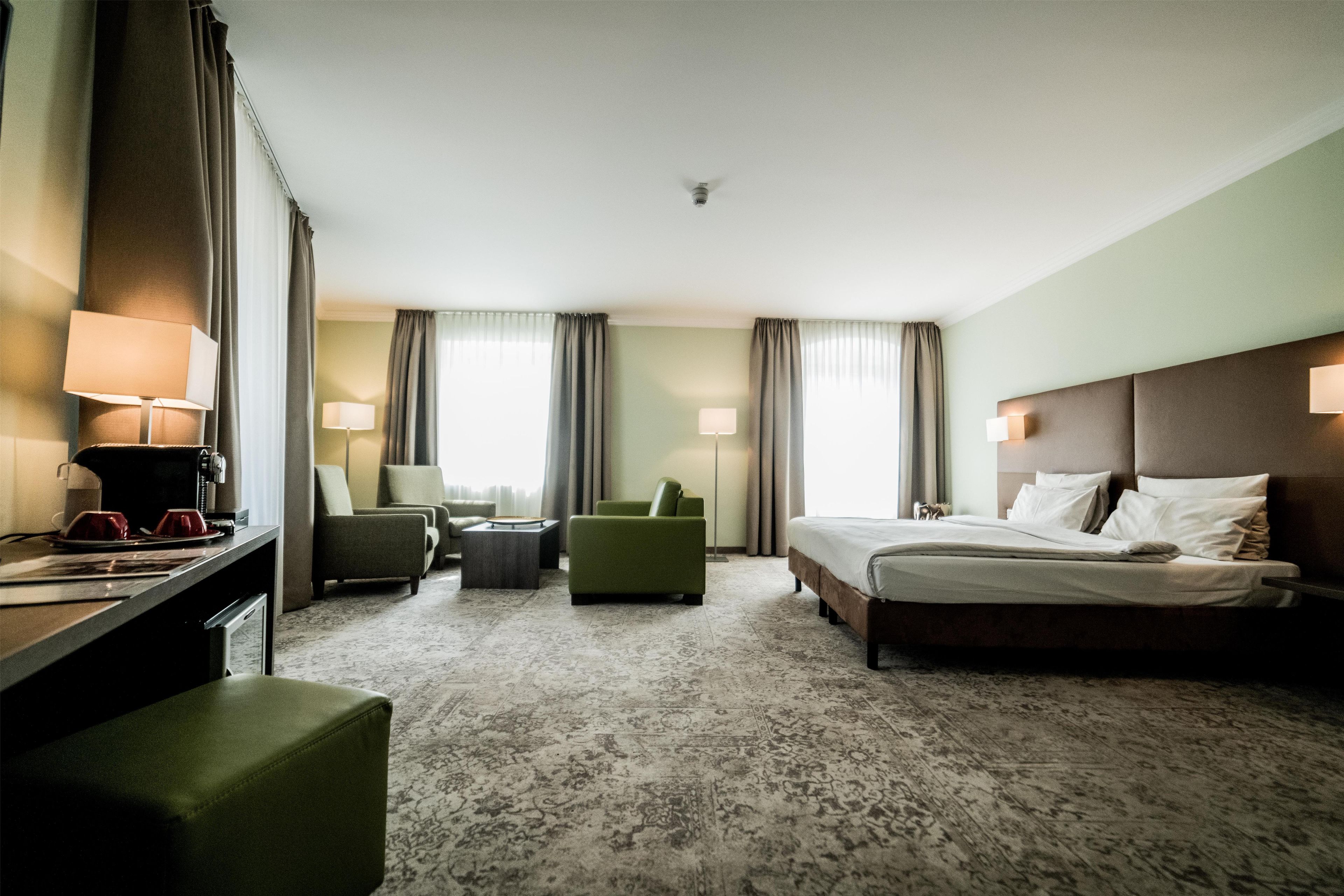 A hotel room with a bed, two armchairs, a small table, a desk with a lamp, and a coffee machine.
