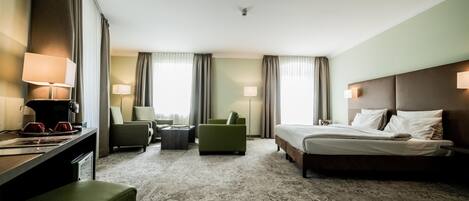 A hotel room with a bed, two armchairs, a small table, a desk with a lamp, and a coffee machine.