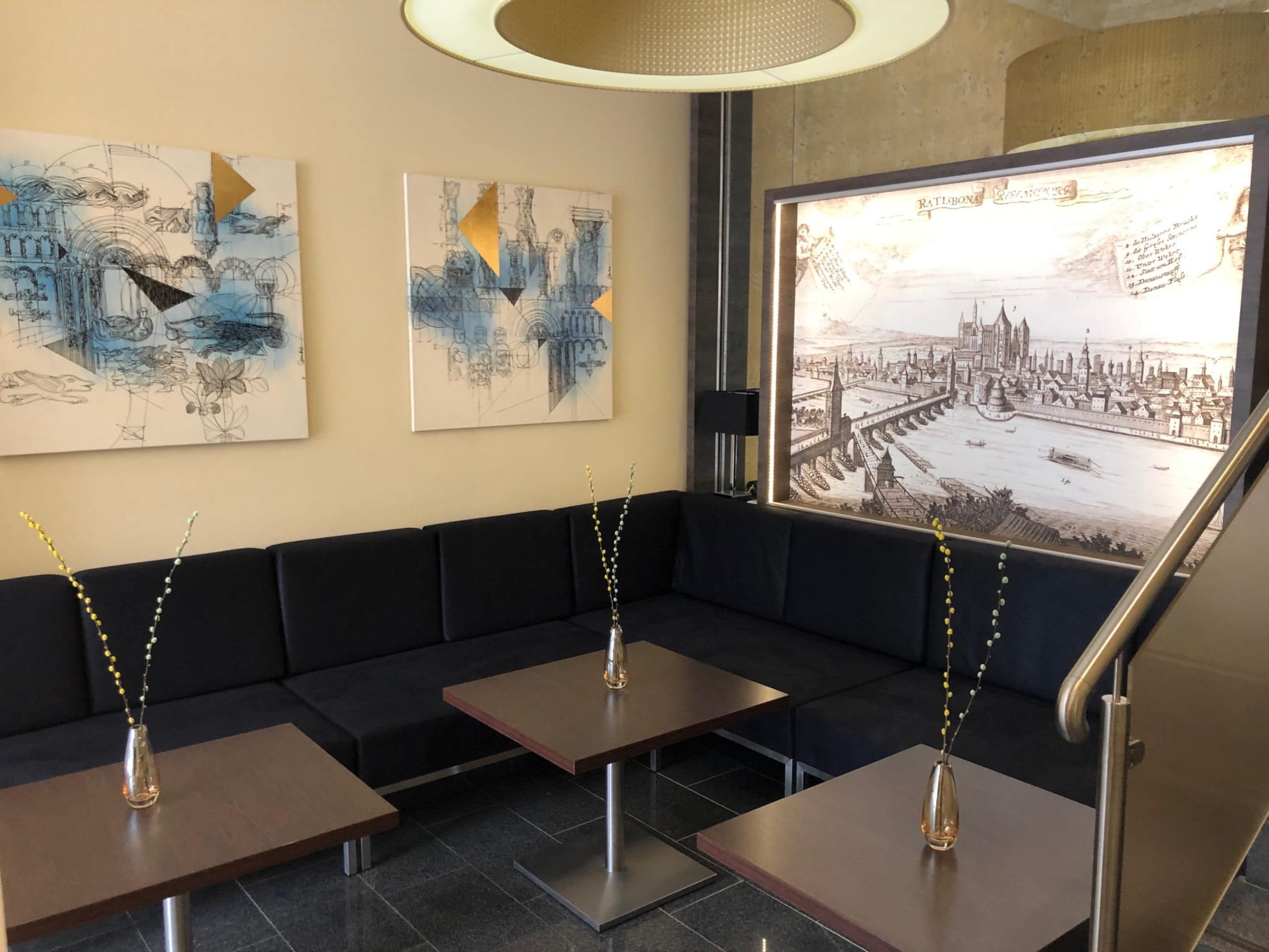 Un intérieur moderne avec un canapé noir, deux tables et une grande œuvre d’art encadrée représentant un paysage urbain.