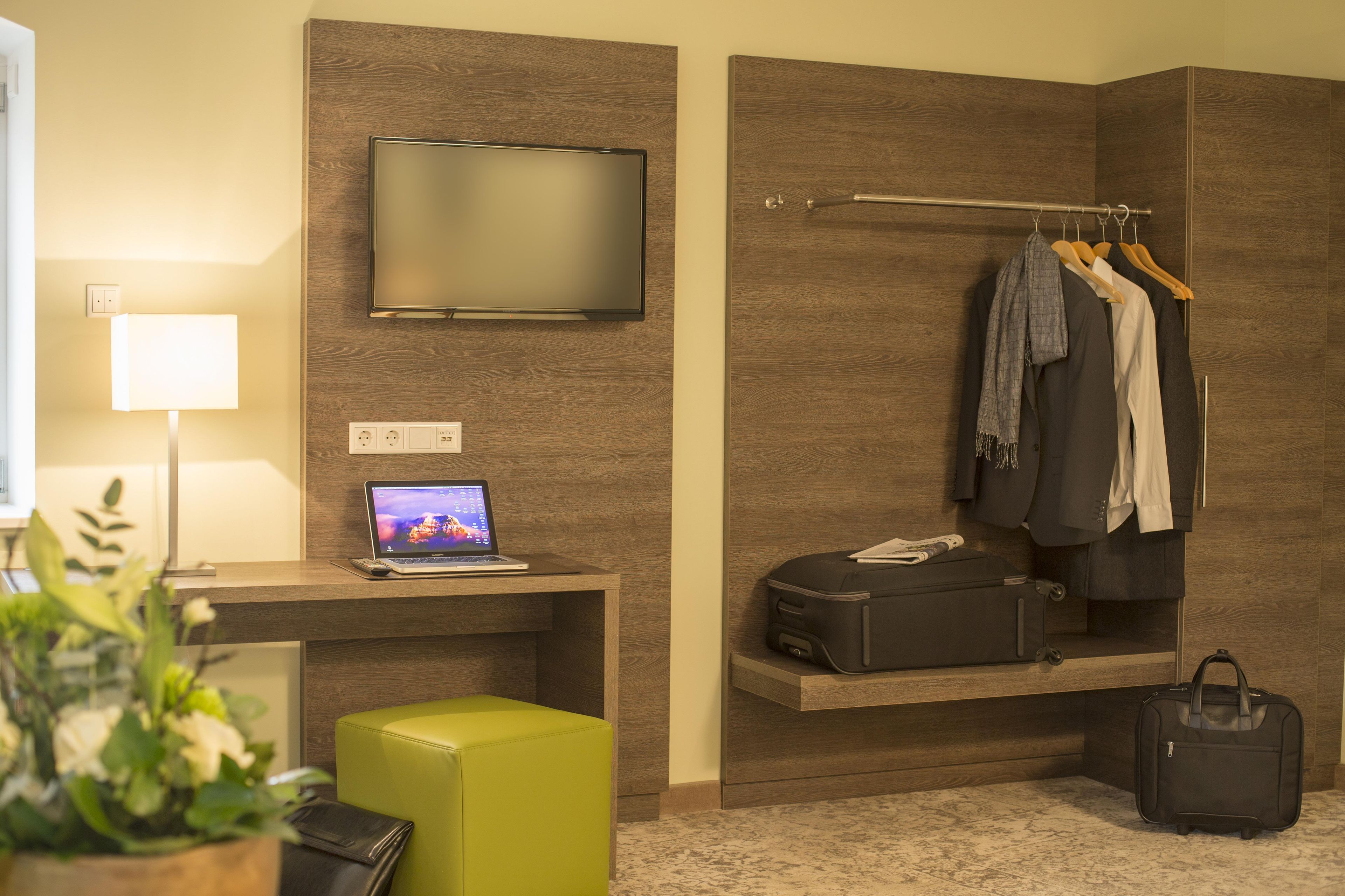 Une chambre d’hôtel équipée d’un téléviseur à écran plat, d’un bureau avec un ordinateur portable, d’un coin salon, d’un porte-manteau avec des vêtements suspendus, d’une valise et d’un petit pouf vert.