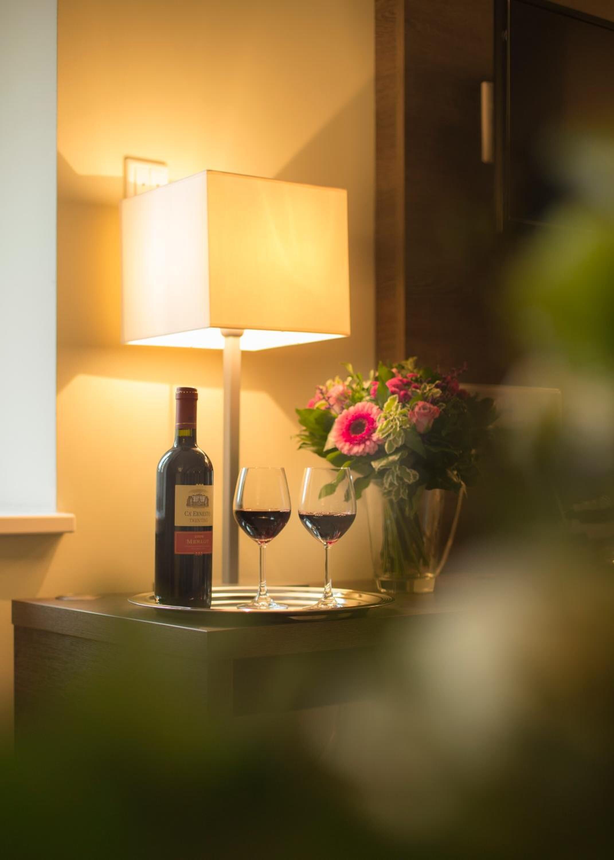 A bottle of wine, two wine glasses, a lamp, and a vase with flowers on a table.