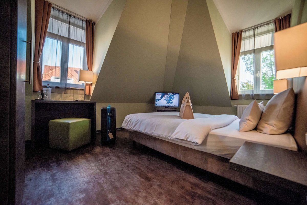 A hotel room with a bed, a desk, a chair, a small table, a TV, and a window with curtains.