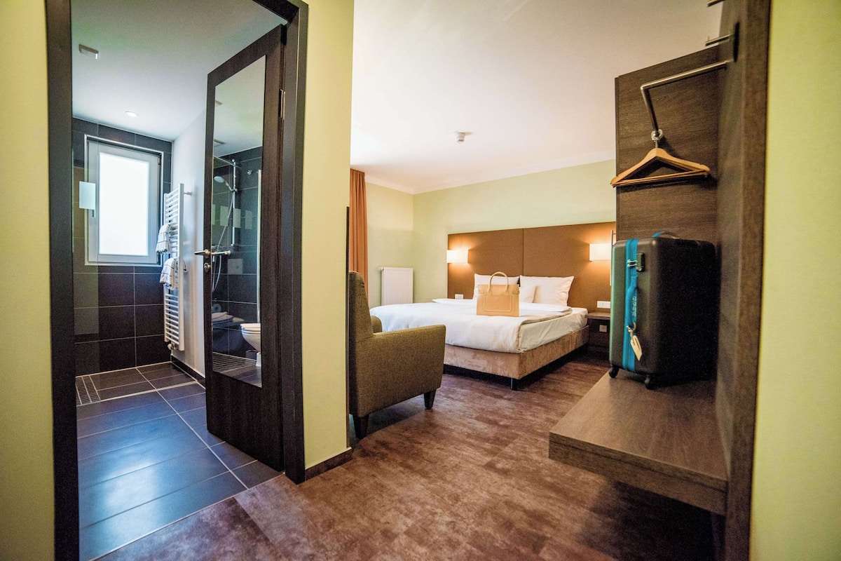 A hotel room with a bed, a chair, a hanging coat rack, and a bathroom with a shower and toilet.