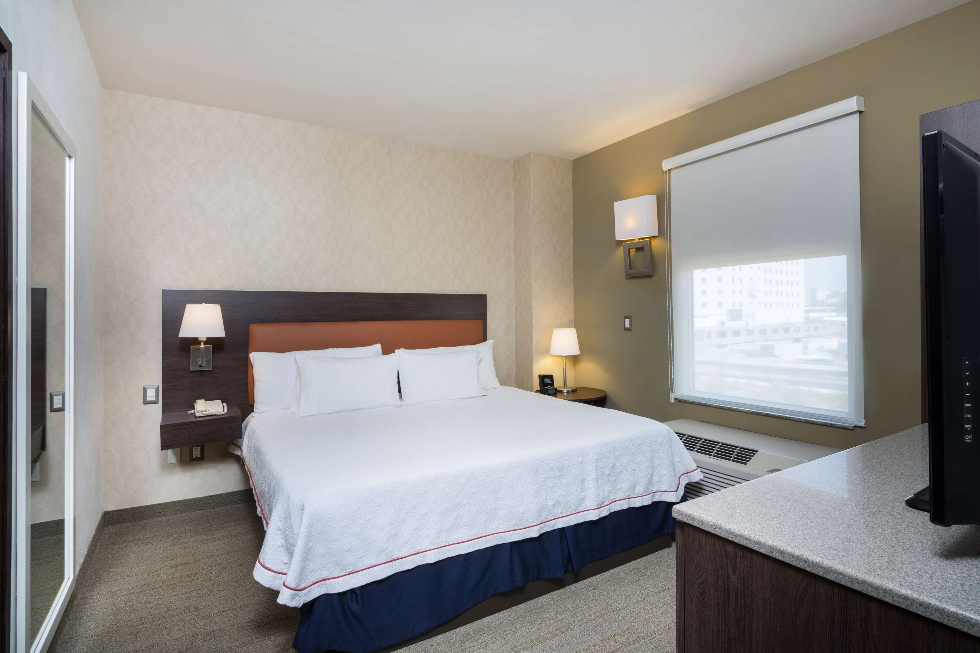 A hotel room with a bed, two bedside lamps, a TV, a window with blinds, and a view of a building.