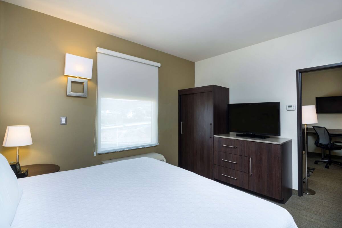 A hotel room with a bed, a TV on a cabinet, a desk with a chair, and a window with blinds.