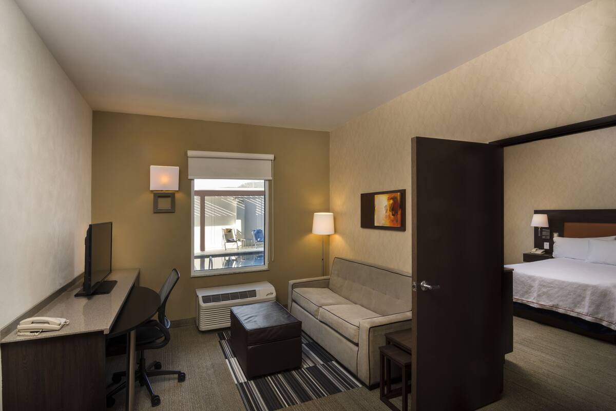 A hotel room with a desk, office chair, sofa, bedside table, lamp, and a bed.