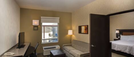A hotel room with a desk, office chair, sofa, bedside table, lamp, and a bed.