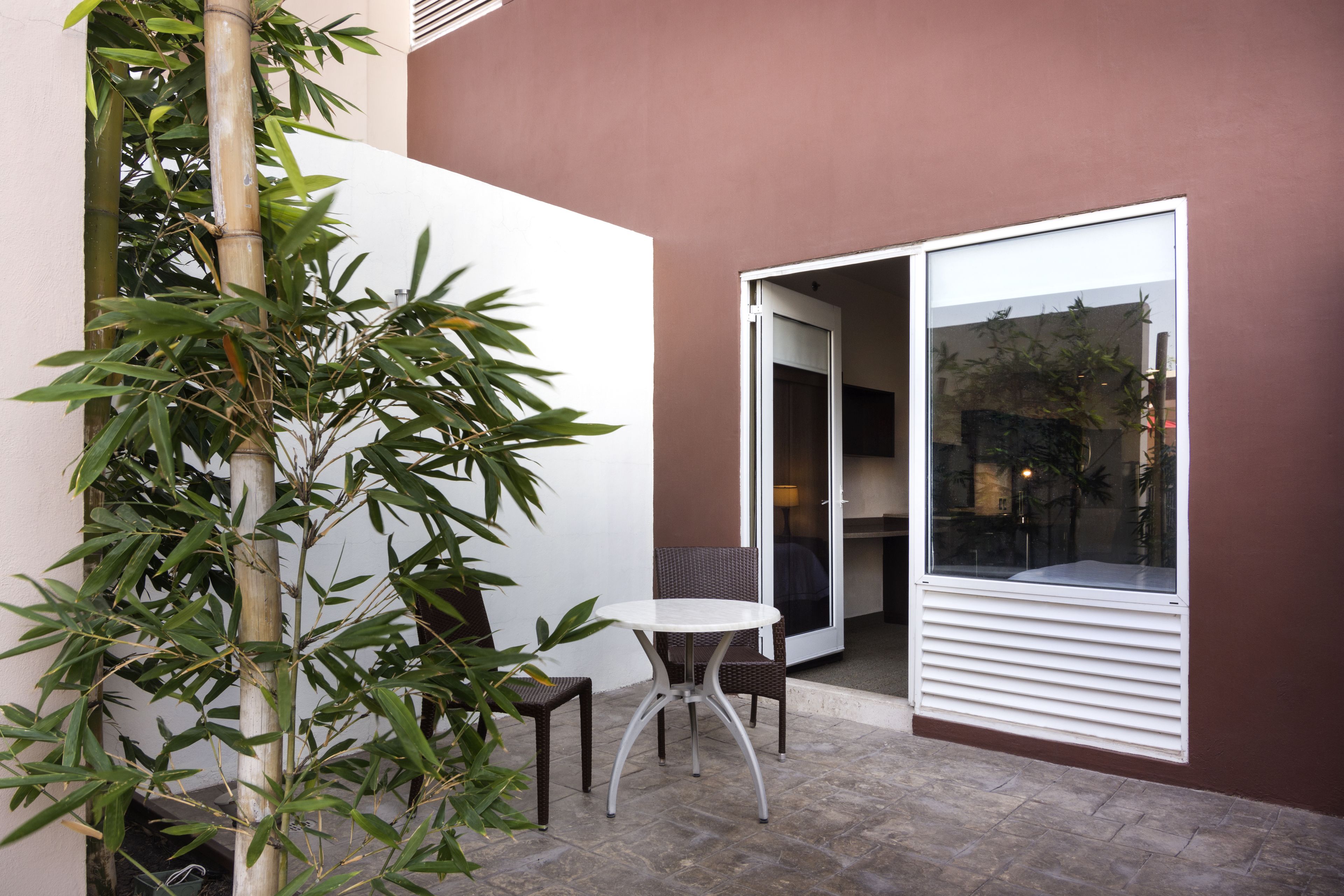 A small outdoor seating area with a table and two chairs, a bamboo plant, and a sliding glass door leading to an indoor space.