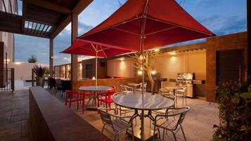 Un patio al aire libre con mesas y sillas redondas, una sombrilla roja y una parrilla.