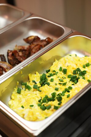 Un bufé con huevos revueltos adornados con cebollines y una sección aparte con tocino.