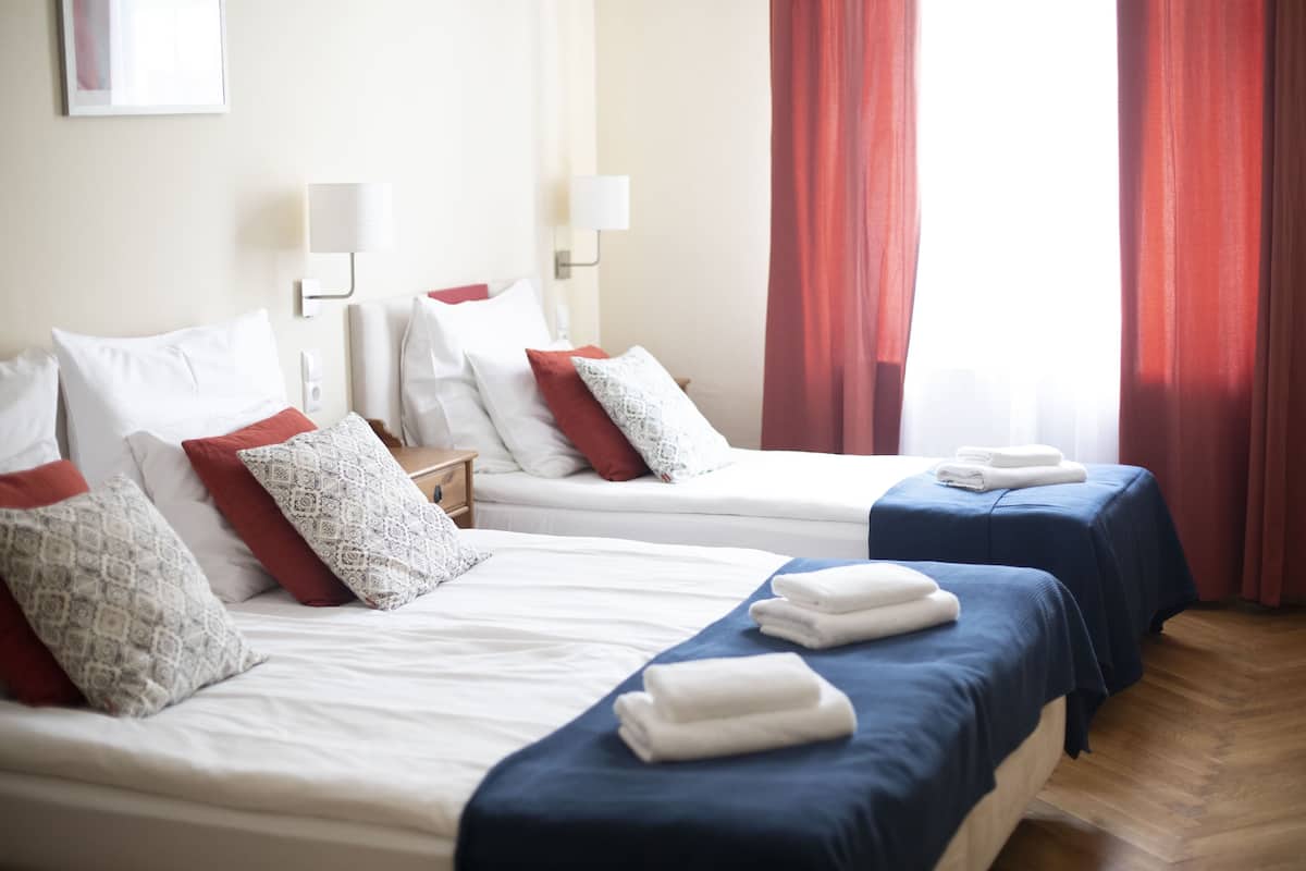 Une chambre d’hôtel avec deux lits, chacun agrémenté de draps blancs et d’oreillers rouges, une table de chevet avec une lampe et une fenêtre avec des rideaux rouges.