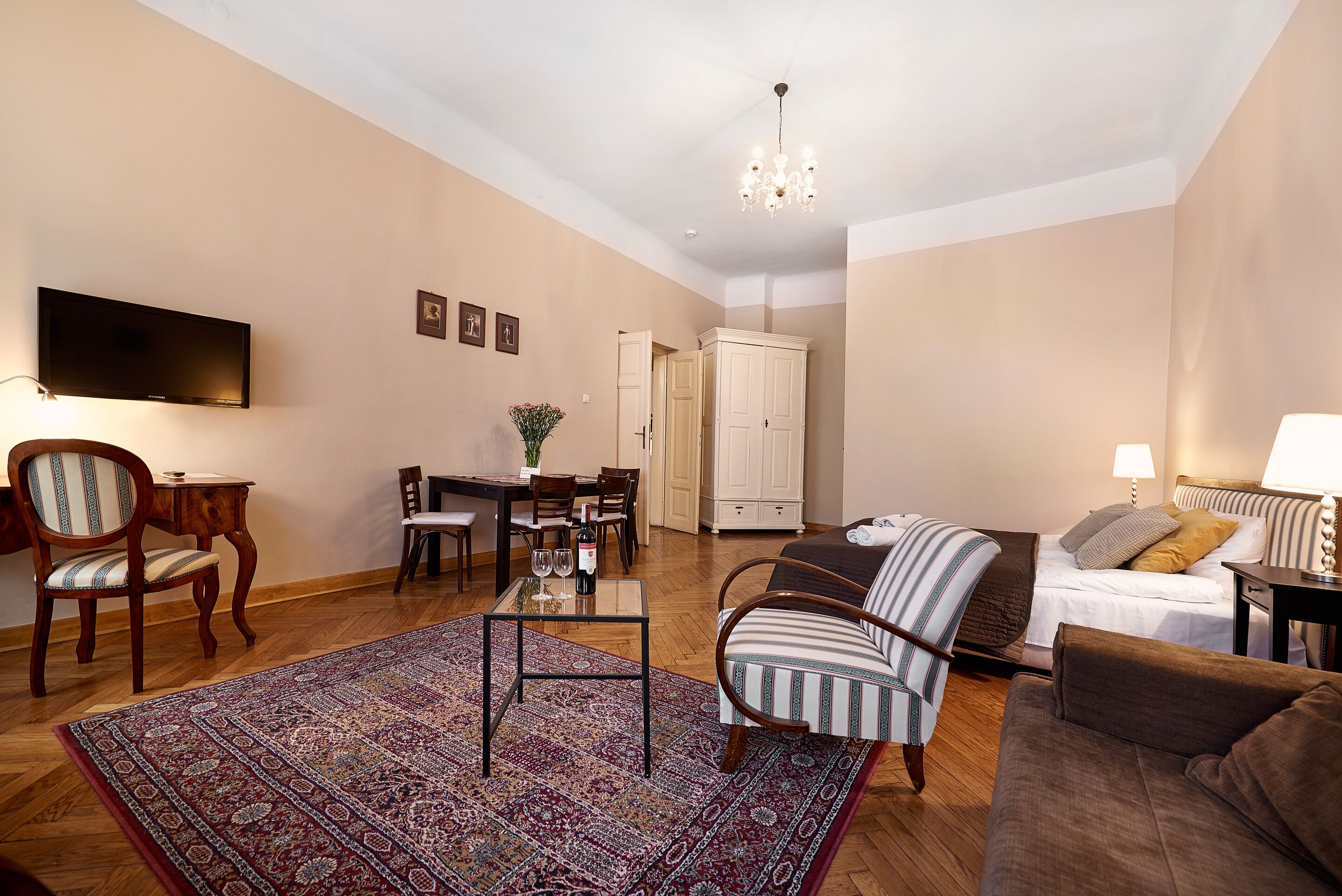 A living room with a television, a dining area with a table and chairs, a sofa, a patterned rug, and a chandelier.