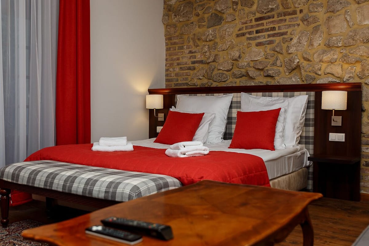 Une chambre d’hôtel avec un mur en pierre, un lit avec une literie rouge et blanche, un plaid et une table de chevet en bois avec une lampe.