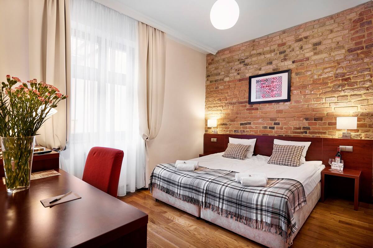 Une chambre d’hôtel avec un mur en briques, un grand lit recouvert d’une couverture à carreaux, un bureau agrémenté d’un vase de fleurs et une fenêtre avec des rideaux.