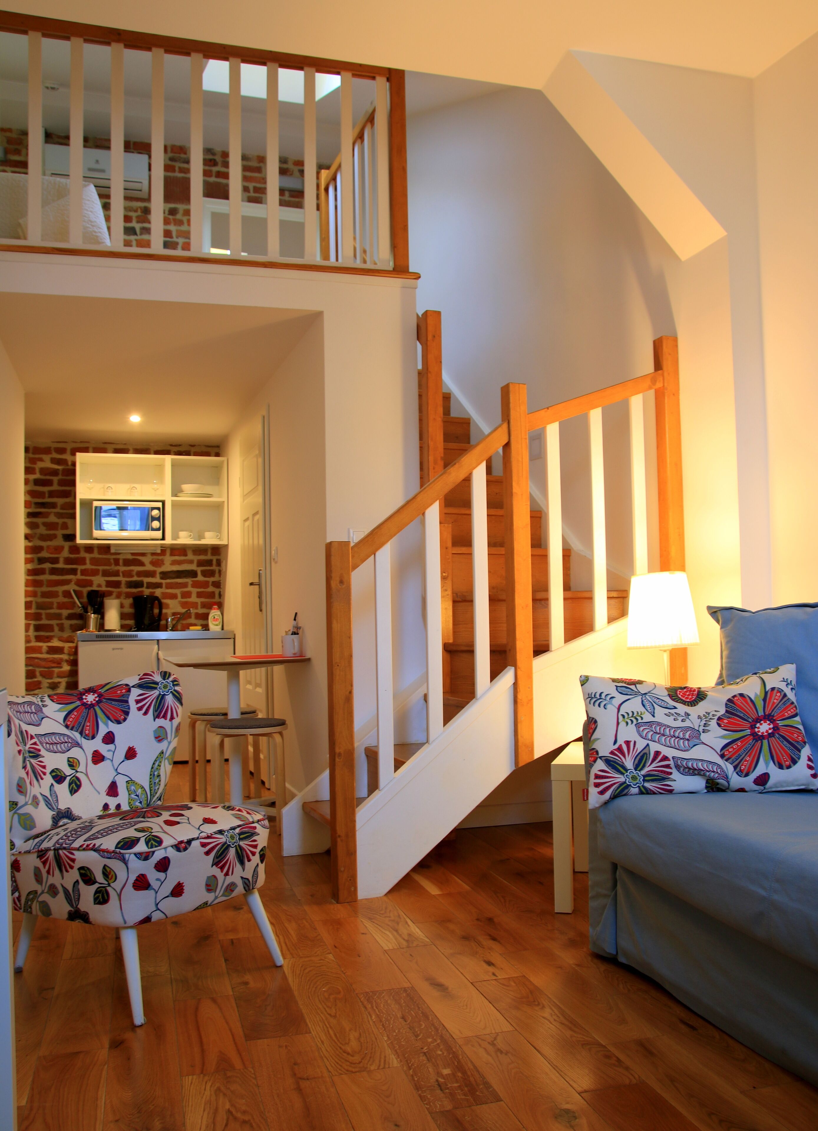 A cozy living room with a wooden staircase, a dining area with a brick wall, and a kitchen with a microwave.