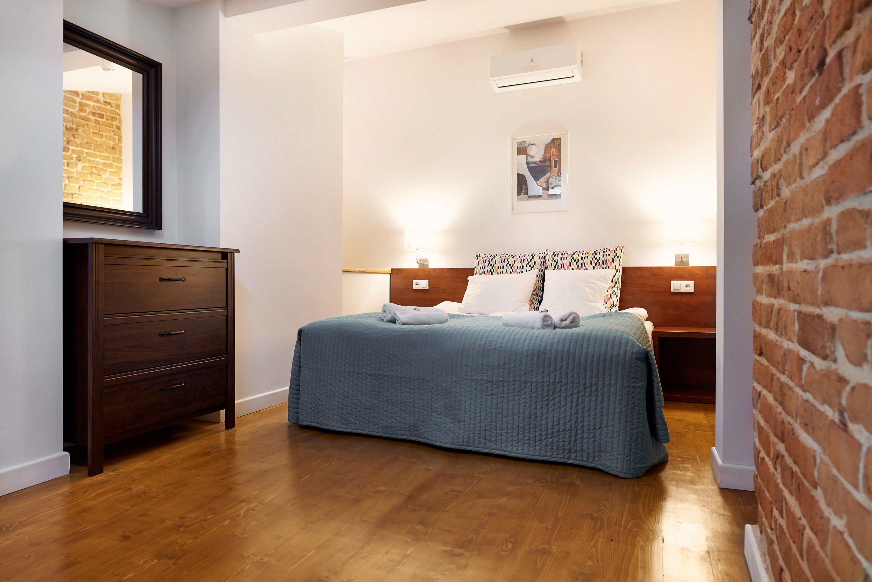 A bedroom with a wooden floor, a bed with a blue quilt, a wooden dresser, a brick wall, and a framed picture on the wall.