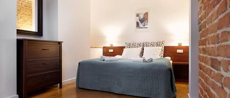 A bedroom with a wooden floor, a bed with a blue quilt, a wooden dresser, a brick wall, and a framed picture on the wall.