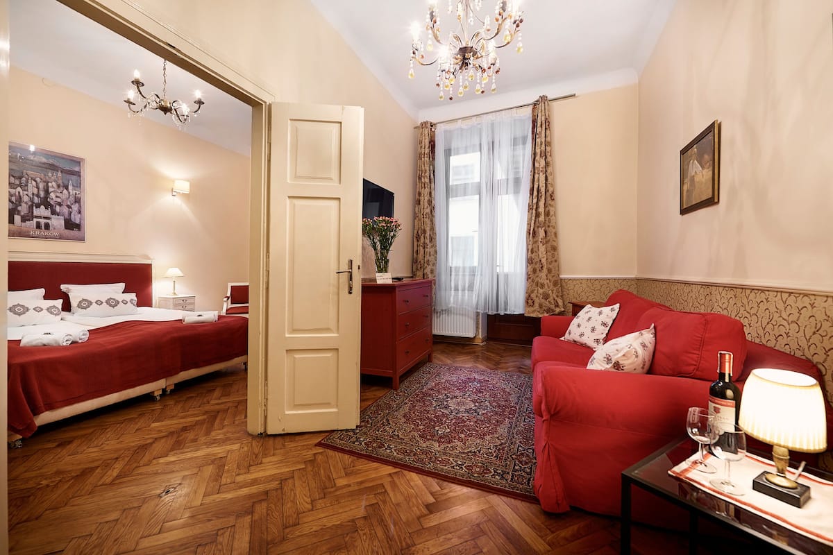 Une chambre d’hôtel avec deux lits, un canapé rouge, une petite table avec une lampe, un lustre et une grande fenêtre avec des rideaux légers.