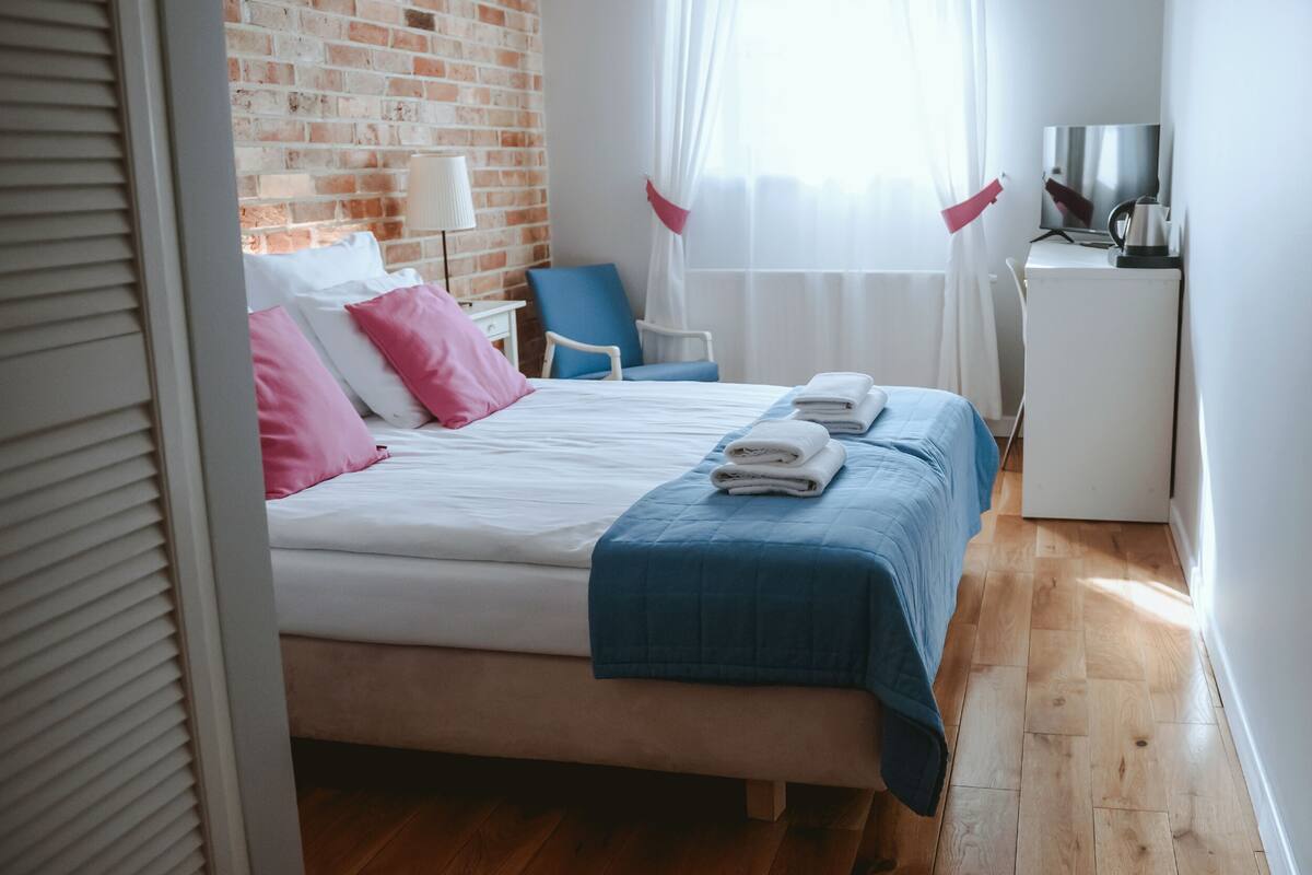 Un lit bien fait, avec une literie rose et bleue, un petit bureau avec une bouilloire et un téléviseur posé sur une commode blanche.