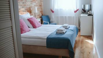Una cama bien hecha con ropa de cama rosa y azul, un pequeño escritorio con una tetera y un televisor sobre un mueble blanco.