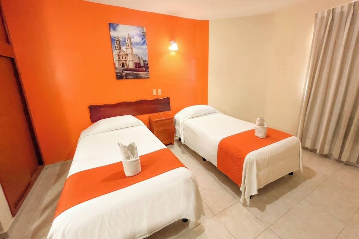Habitación de hotel con dos camas, paredes anaranjadas, un cuadro de una catedral y una ventana con cortinas.