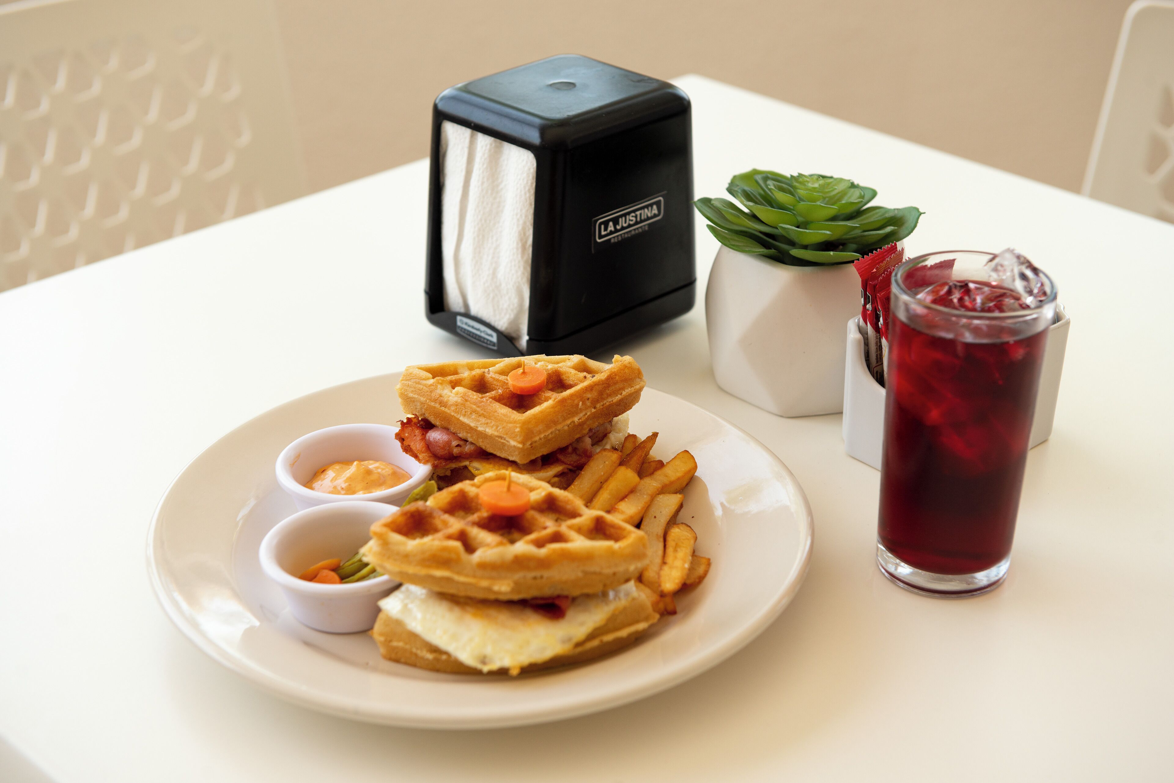 A plate with waffles, fries, and a drink, along with a waffle maker and a plant on a table.