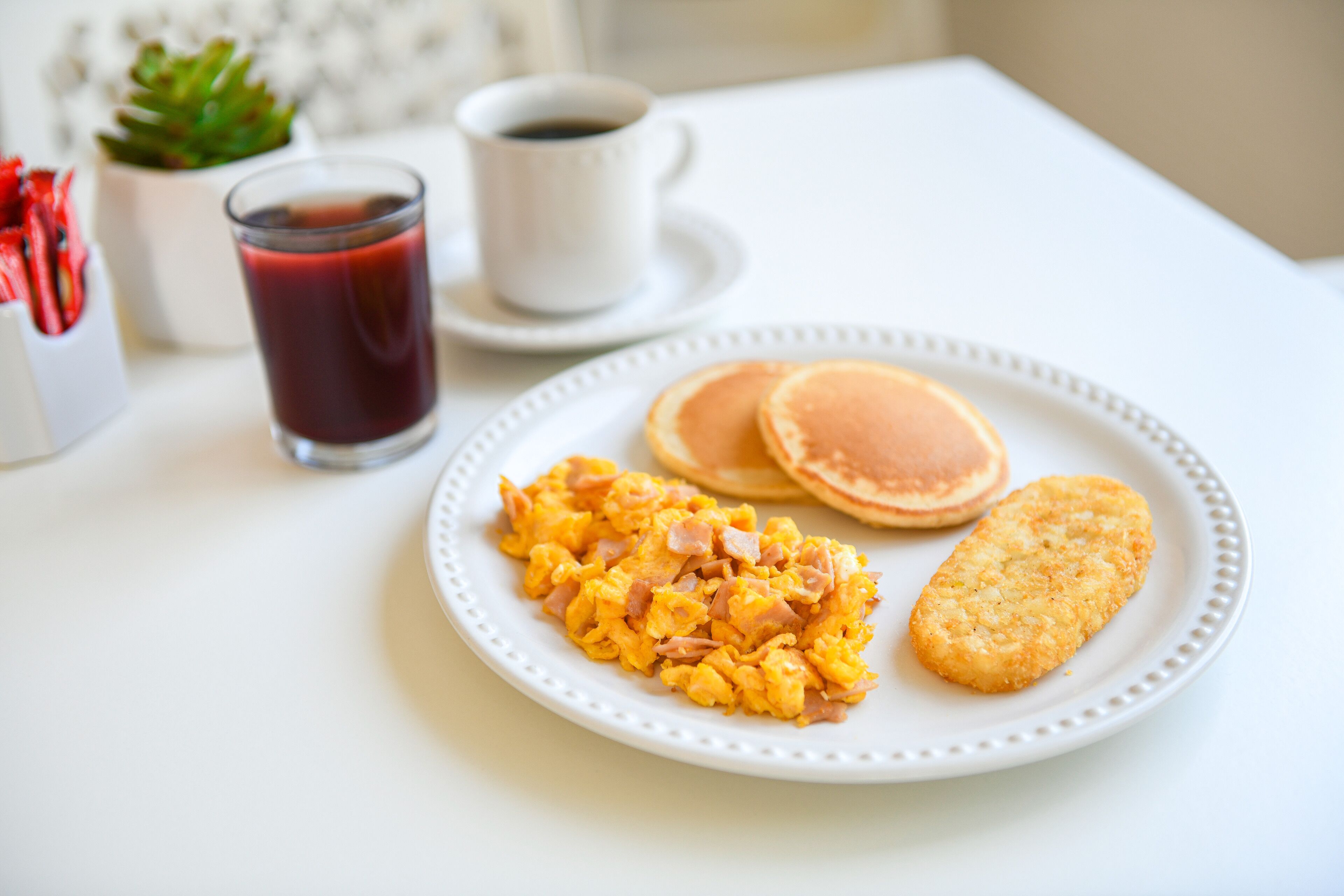 A breakfast plate with scrambled eggs, bacon, and a hash brown, accompanied by a glass of red juice and a cup of coffee on a white table.