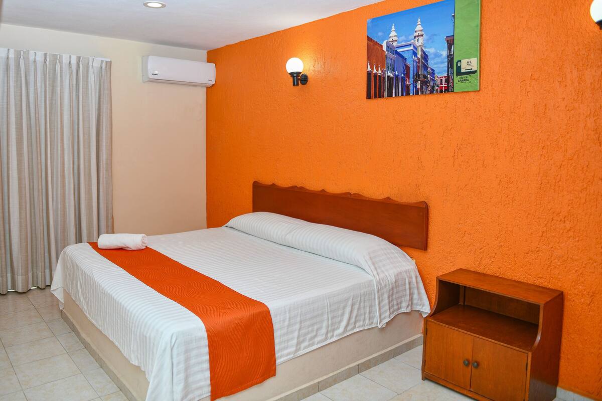 Habitación de hotel con cama, mesita de noche, equipo de aire acondicionado y lámpara fijada a la pared.