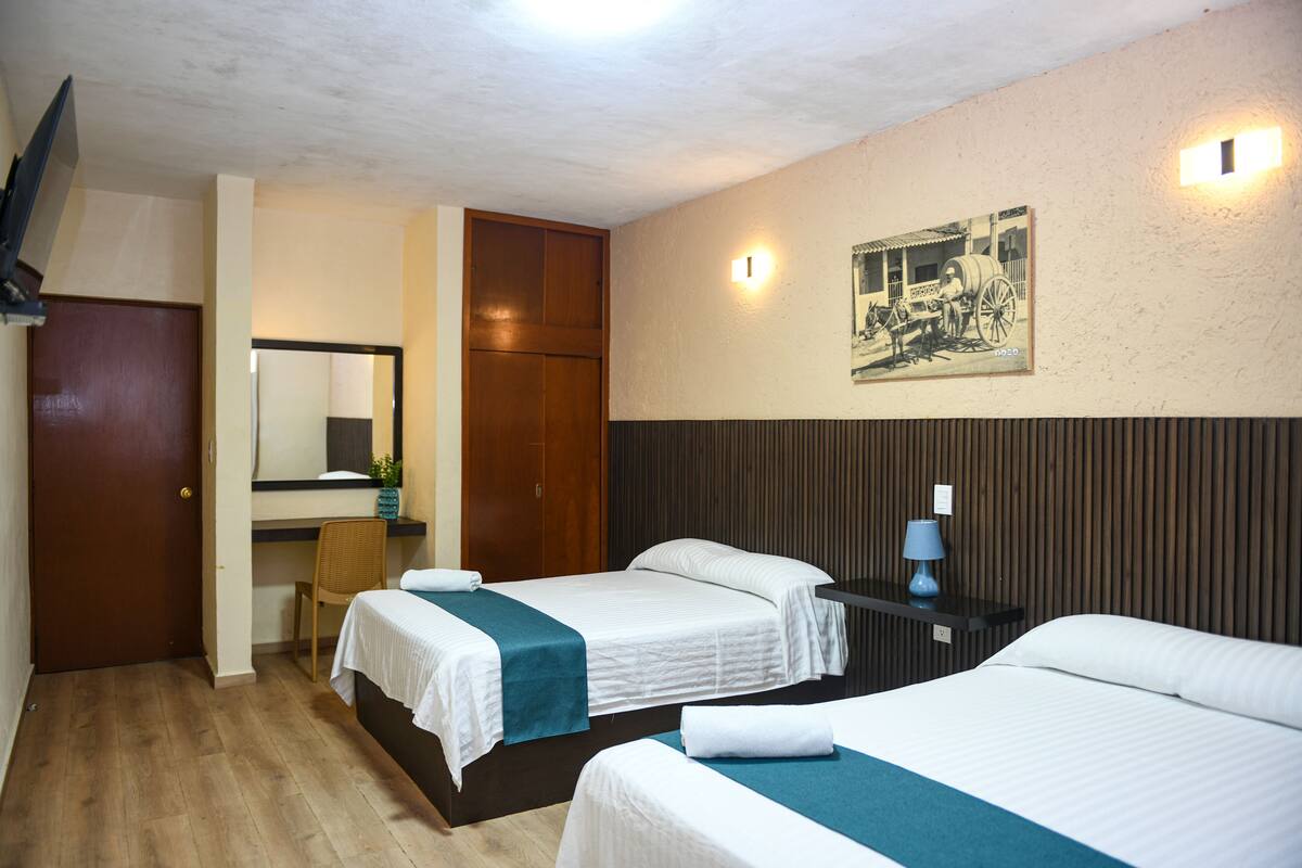 Habitación de hotel con dos camas, un armario de madera, un escritorio con silla y un cuadro colgado en la pared.