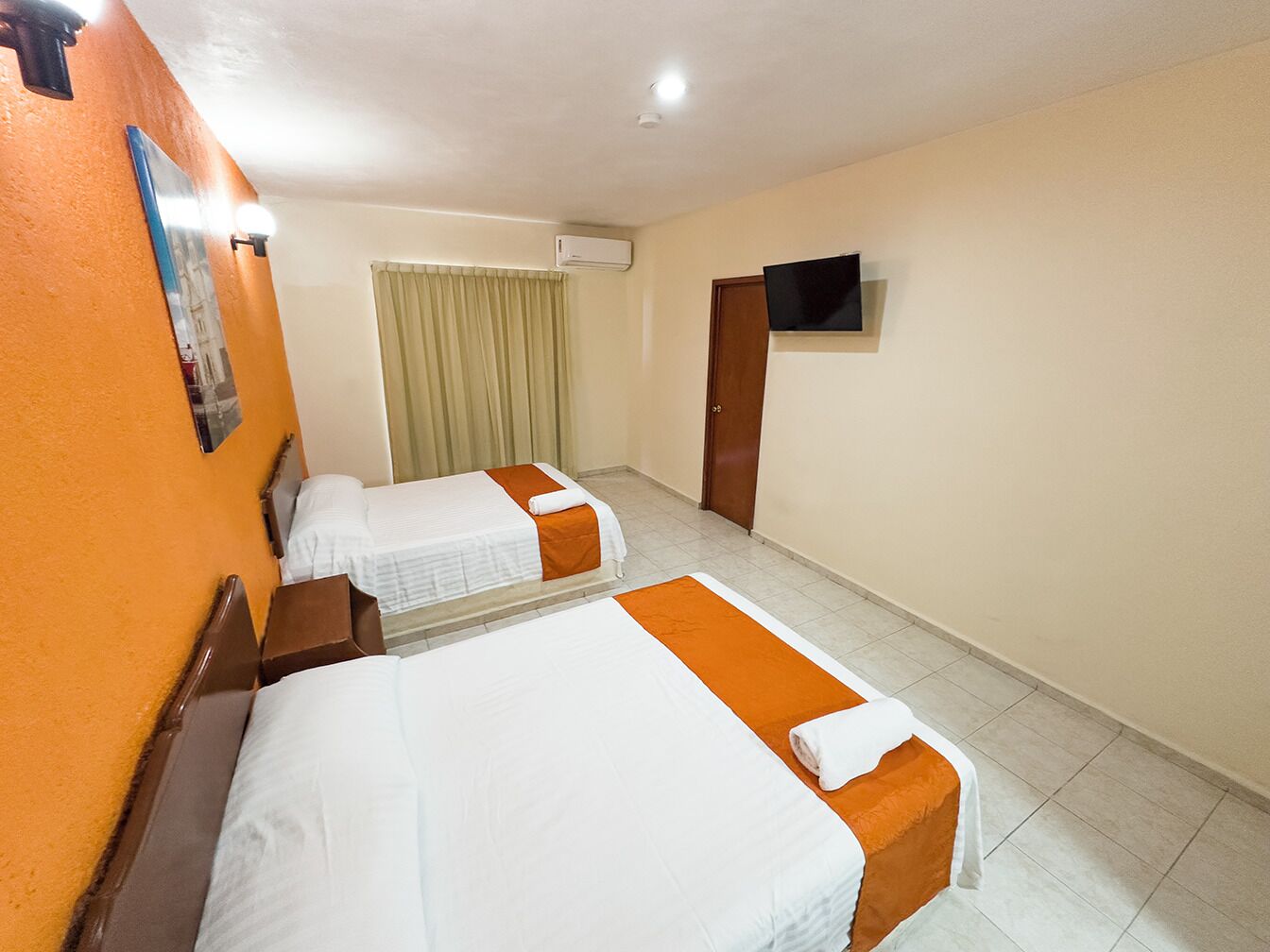 A hotel room with two beds, a television, an air conditioning unit, and a window with curtains.