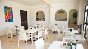 A bright, spacious dining area with white tables and chairs, a ceiling fan, and colorful artwork on the walls. - Hotel U Xul Kah (Campeche)