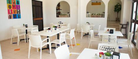 Un comedor amplio y luminoso con mesas y sillas blancas, un ventilador de techo y coloridas obras de arte en las paredes.
