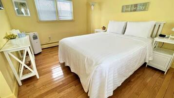 A bedroom with a large bed, a bedside table, a nightstand, a lamp, and a window with blinds.