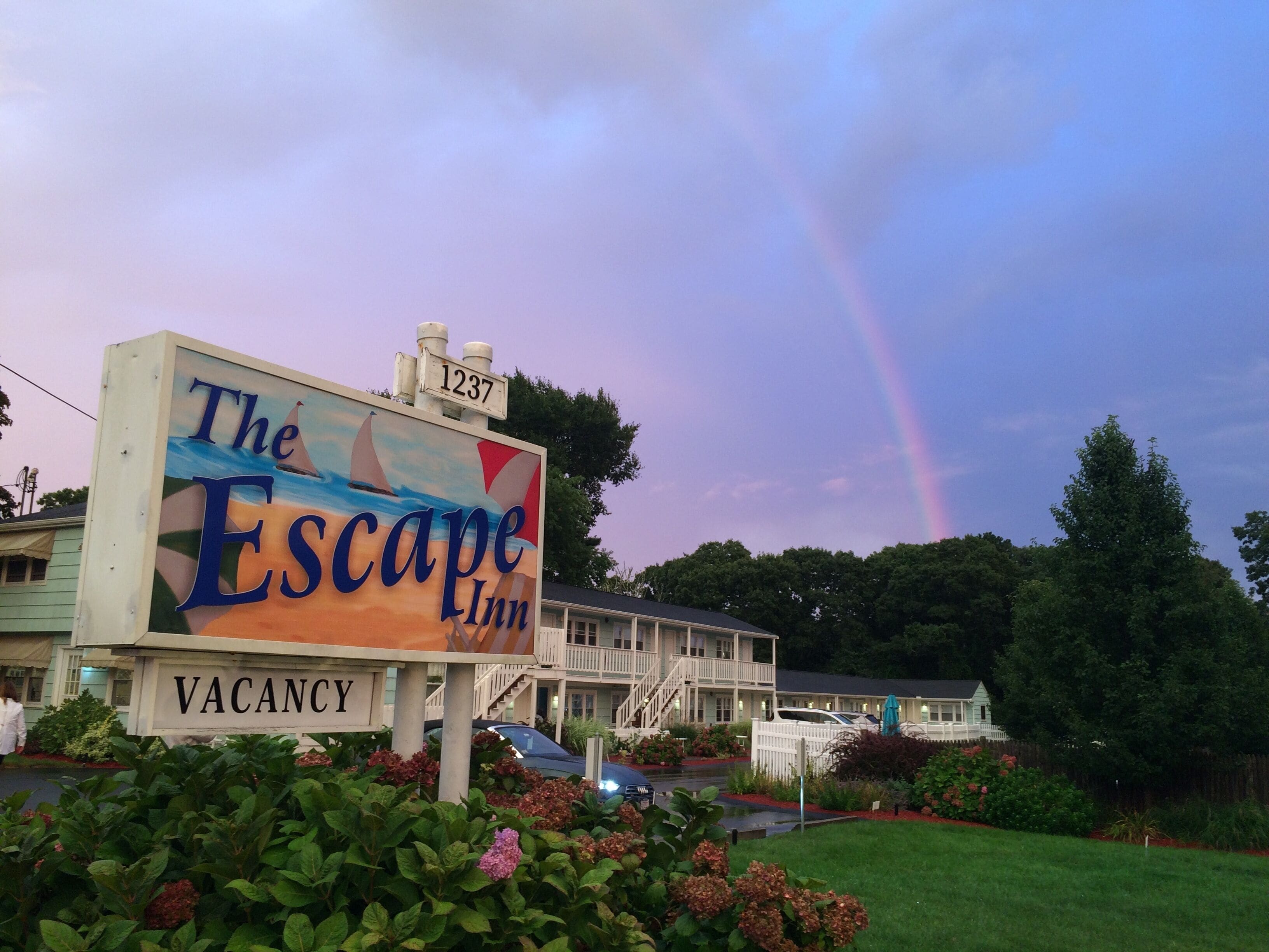 A sign for The Escape Inn with a 'VACANCY' label, surrounded by lush greenery and a rainbow in the sky.