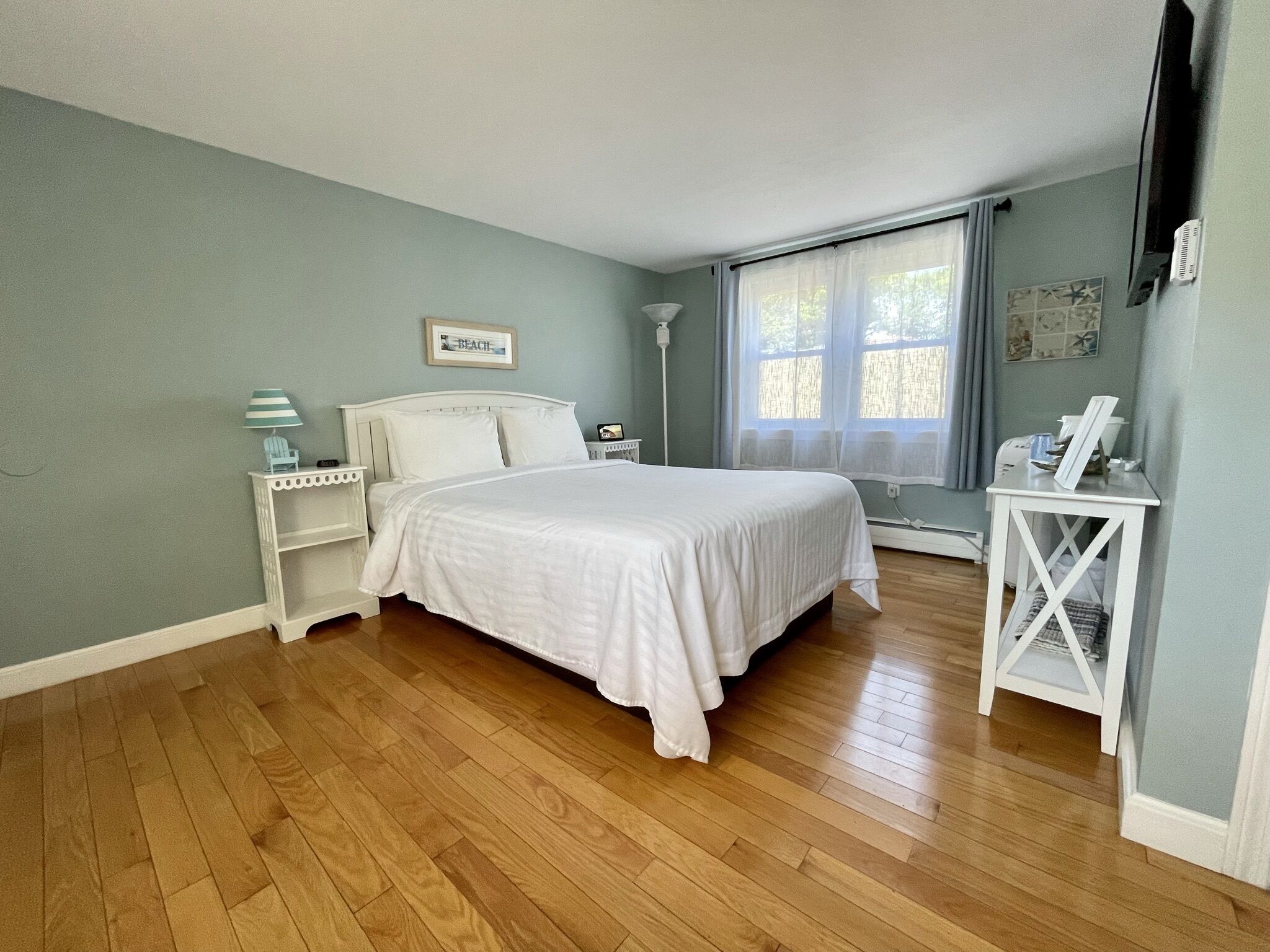 A bedroom with a bed, nightstands, a lamp, a window with curtains, and a small table with a picture frame.