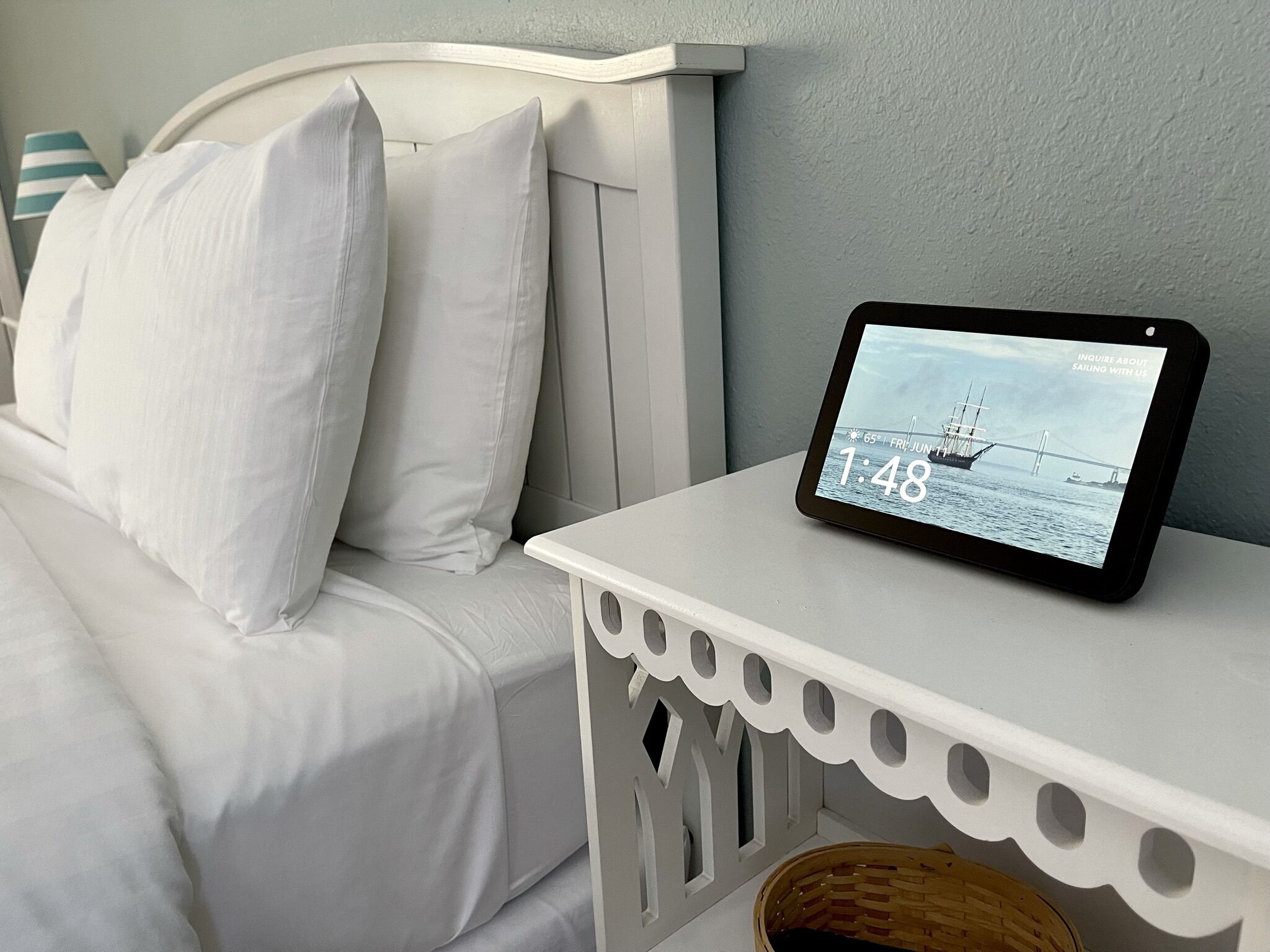 A white bed with pillows, a tablet displaying a bridge and ship, and a white nightstand with a basket.