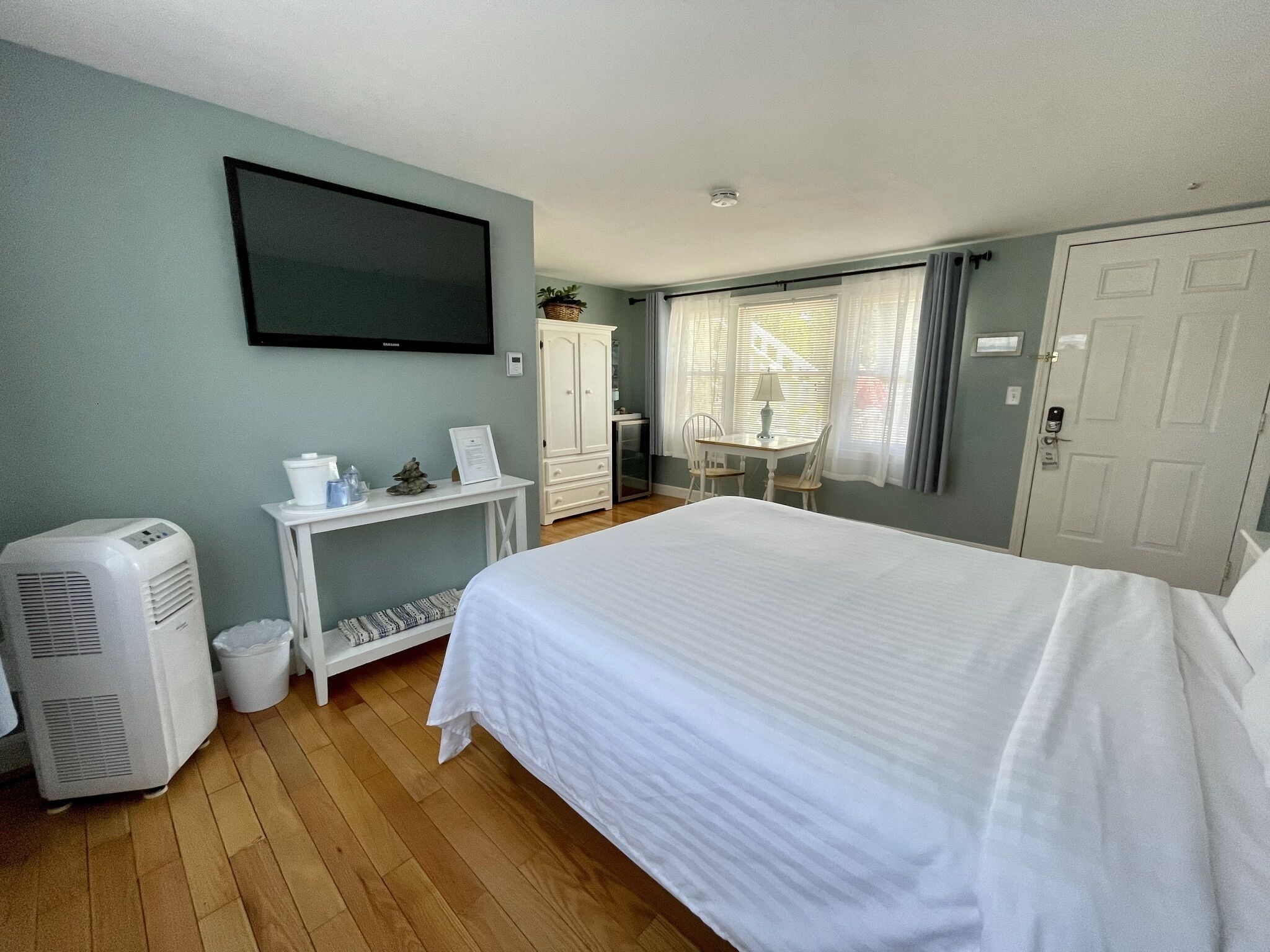 A hotel room with a bed, a TV mounted on the wall, a desk with a chair, a wardrobe, and a window with curtains.