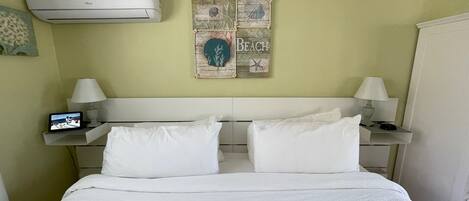 Quarto de hotel com cama, duas mesas de cabeceira com luminárias, ar condicionado fixado na parede e um objeto decorativo na parede.