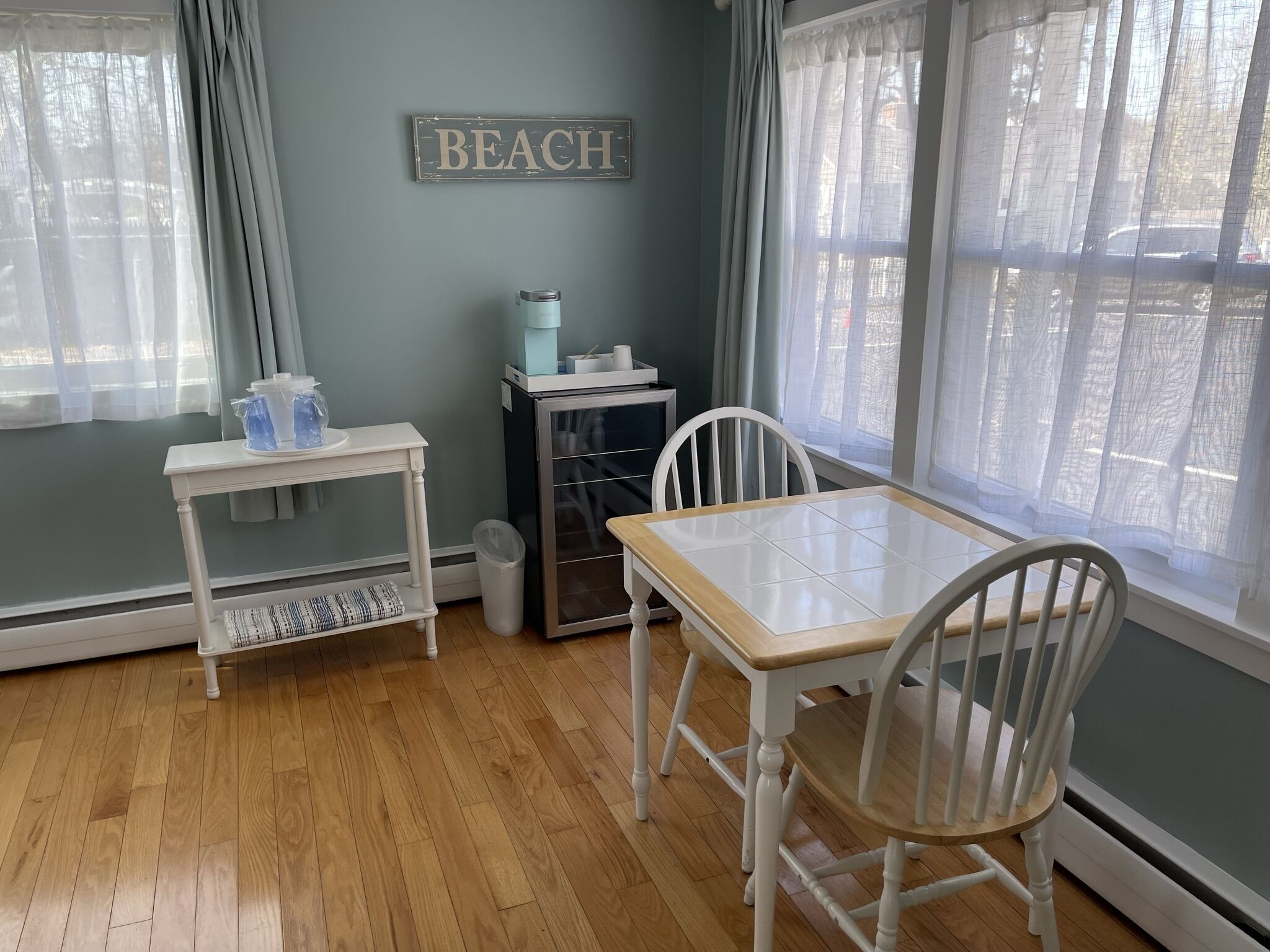 Una habitación con piso de madera, una mesa blanca con dos sillas, una mesita blanca con dispensador de agua, un bote de basura y un cartel en la pared que dice "BEACH".