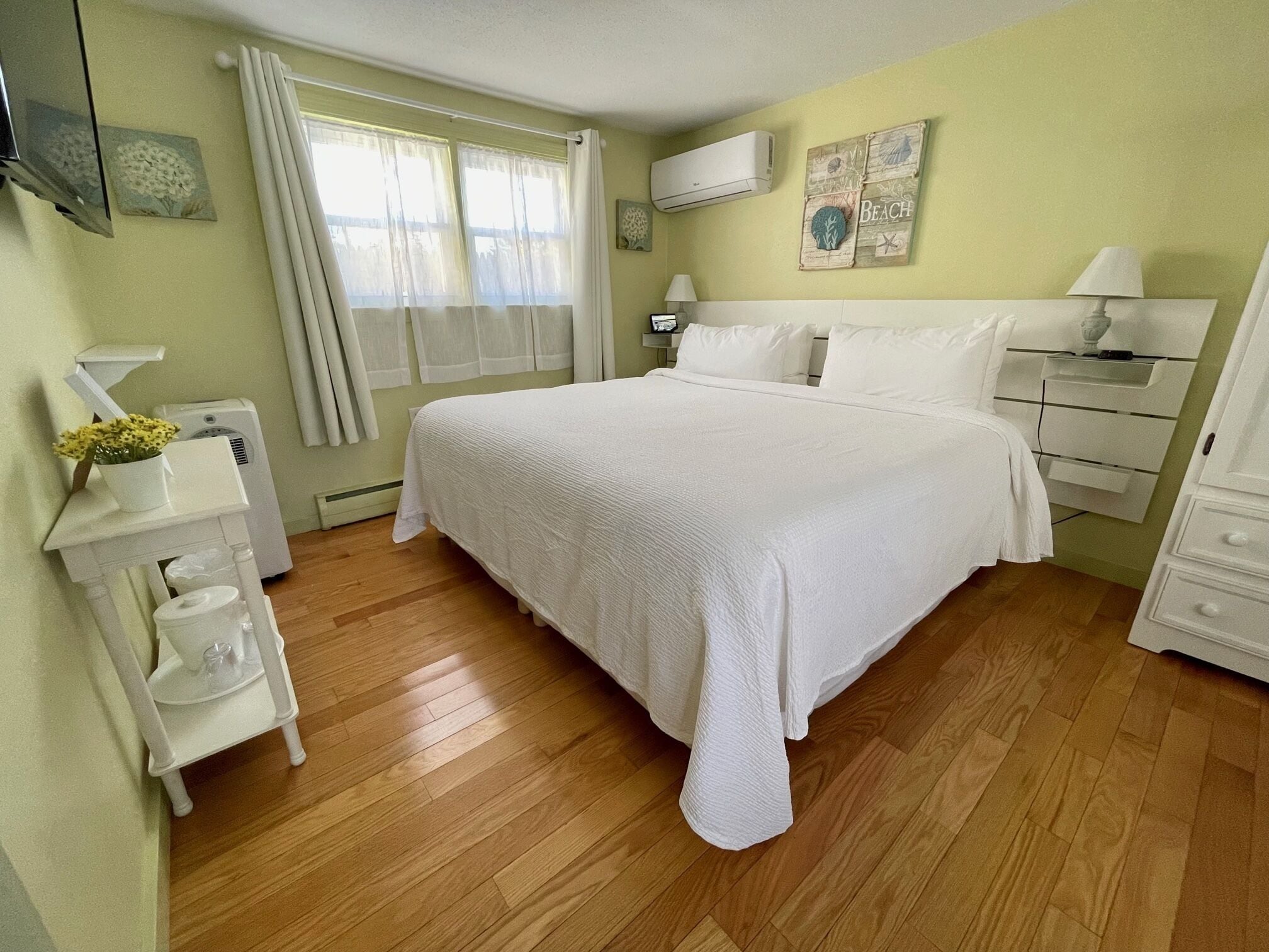 A bedroom with a large bed, bedside tables, a nightstand, a lamp, a wall-mounted air conditioner, and a window with curtains.