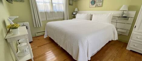 A bedroom with a large bed, bedside tables, a nightstand, a lamp, a wall-mounted air conditioner, and a window with curtains.
