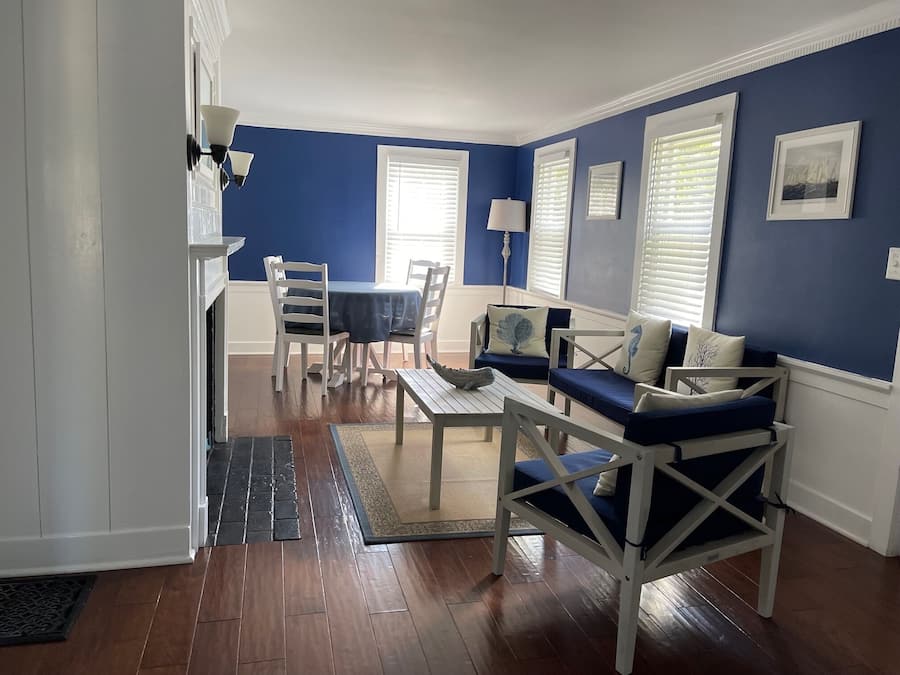 A living room with a blue wall, wooden flooring, a dining table with chairs, a sofa with cushions, and a lamp.