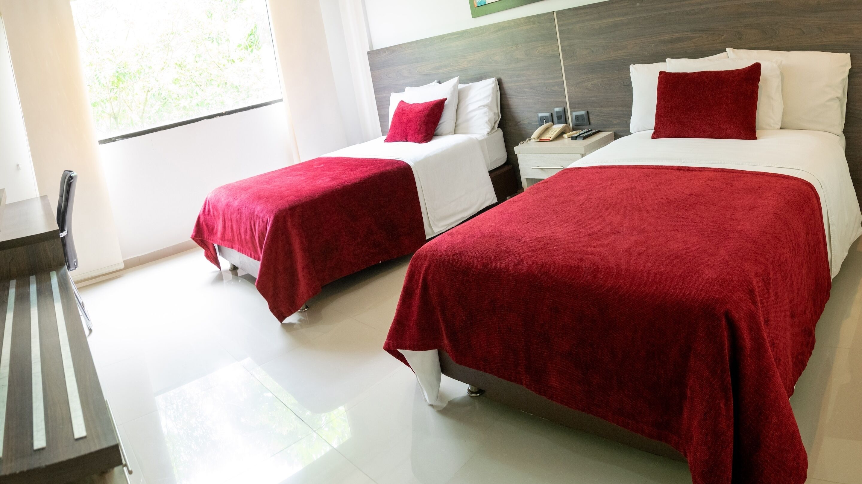 Habitación de hotel con dos camas, cada una con colcha roja y almohadas blancas. Entre las camas, una mesita de noche con un teléfono y una lámpara.