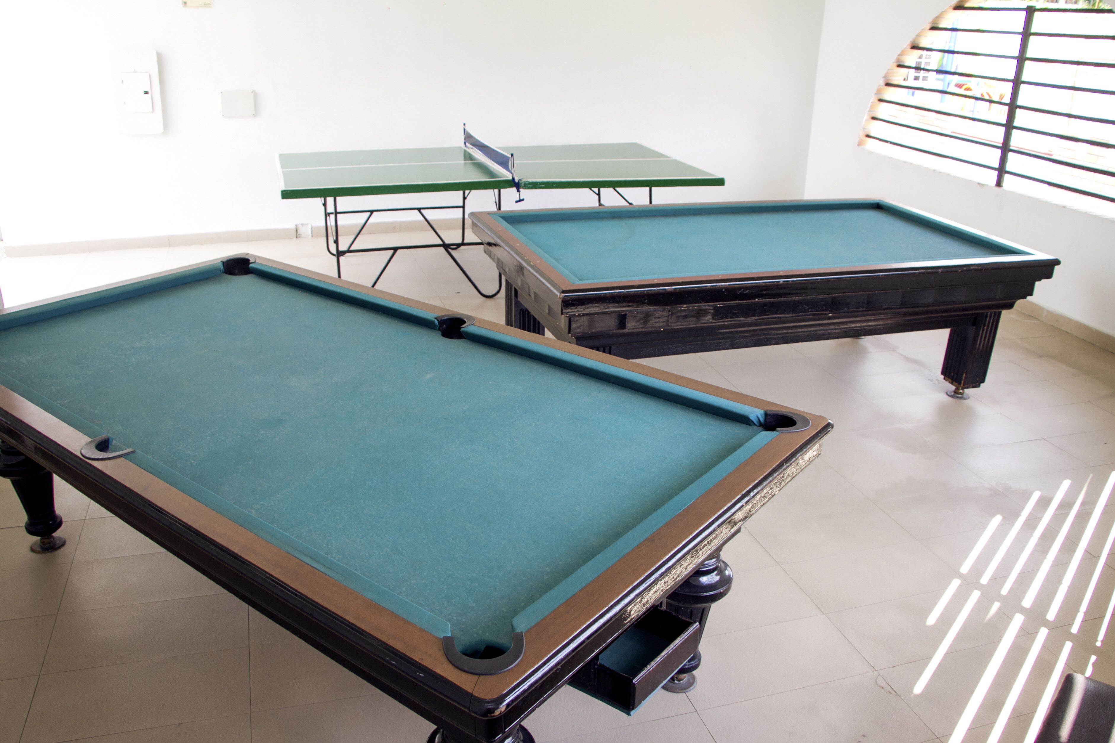 Two pool tables in a room with white walls and a window with blinds.