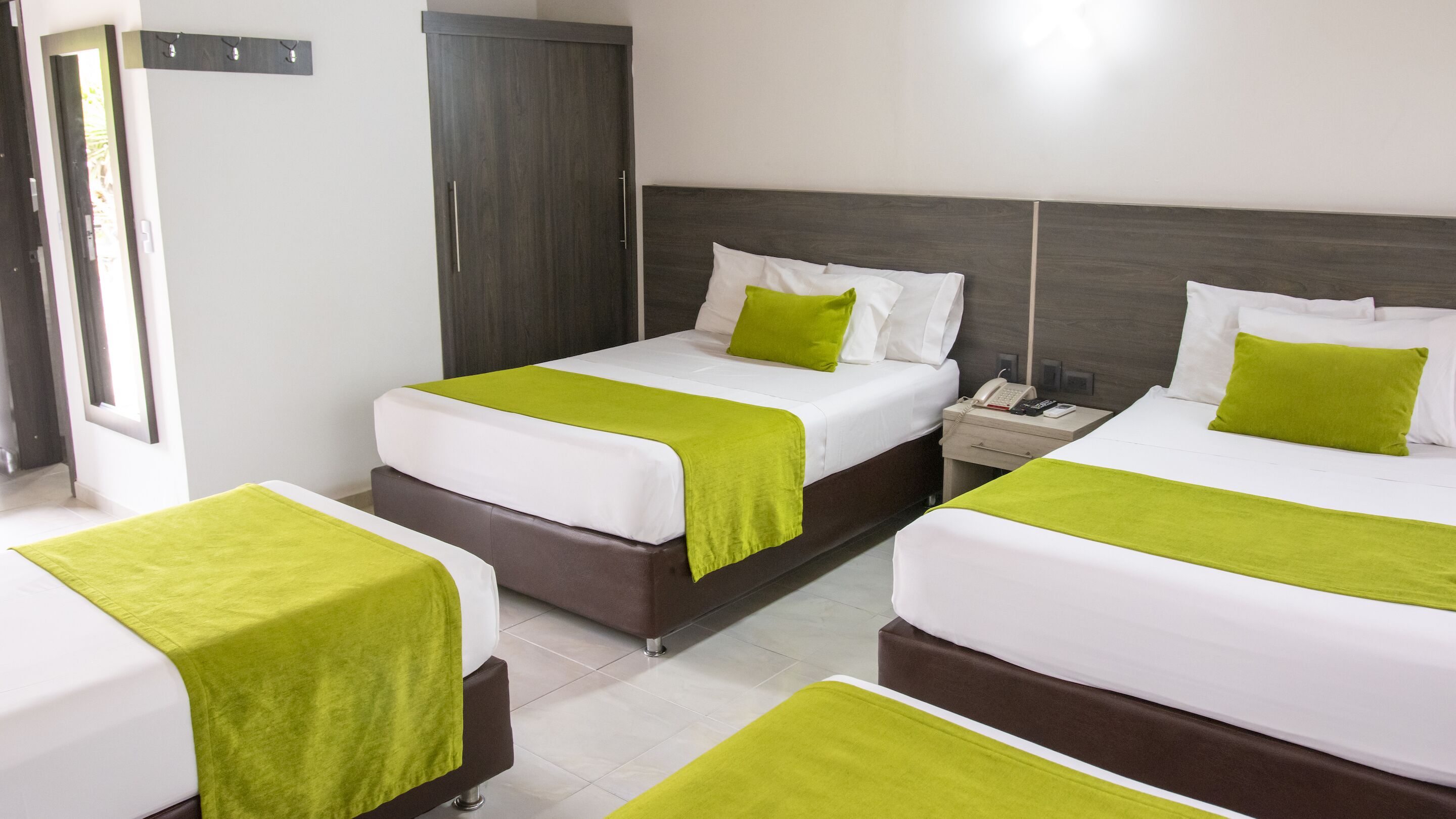 Habitación de hotel con dos camas, cada una con una manta verde y ropa de cama blanca. Hay una mesita de noche con un teléfono y una lámpara.